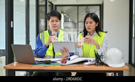 Due ingegneri asiatici professionisti e seri che indossano giubbotti di sicurezza guardano lo schermo di un tablet, discutono di lavoro e lavorano a un progetto in lontananza Foto Stock