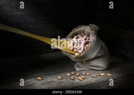 Una natura morta di un mucchio di fagioli Borlotti crudi, secchi, crudi, crudi, beige, bruni in una borsa assiana e cucchiaio di legno nell'angolo di una scatola di legno Foto Stock