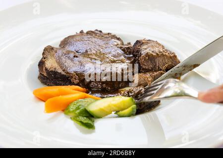Primo piano della bistecca di manzo italiana ben preparata con forchetta e coltello servita su un grande piatto bianco Foto Stock