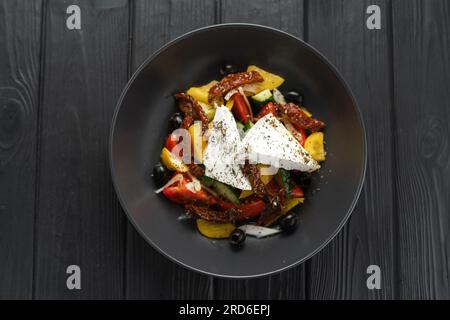 Insalata greca fresca. Piatto con feta, pomodori, cetrioli, pepe rosso, olive nere, pomodori secchi e cipolla su tavola di legno nero. Foto Stock