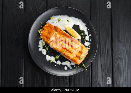 Salmone appena grigliato con fette di limone condite con salsa cremosa su uno sfondo di legno scuro, vista dall'alto Foto Stock
