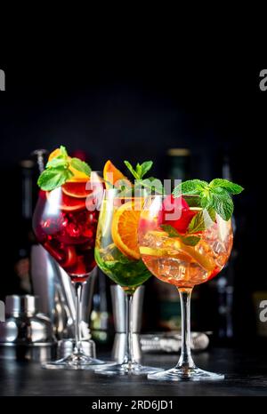 Cocktail alcolici estivi con sangria rosa, rosso, bianco, bevande con vino spagnolo, frutta, agrumi e ghiaccio. Sfondo nero del bancone del bar, utensili da bar in acciaio e. Foto Stock