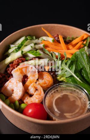 Ciotola con gamberetti, carote, cetrioli, fagioli e verdure con erbe, su sfondo nero. Concetto di cibo sano Foto Stock