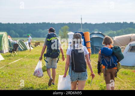 Vista posteriore di turisti irriconoscibili con zaini che trasportano borse mentre cammini lungo le tende da campeggio su prato erboso durante l'evento Foto Stock