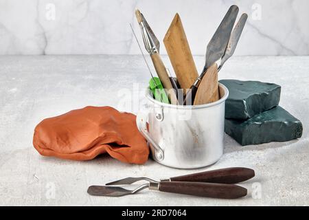 Strumenti di scultura e modellazione in tazza di alluminio circondati da argilla rossa e plasticina verde su un tavolo in pietra chiara. Foto Stock