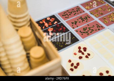 Concetto di cioccolato di alta qualità. Collezione di barrette di cioccolato fatte a mano accanto ai coni di gelato per waffle. Cioccolato bianco rubino e fondente in stampi decorati con lamponi pistacchios noci frutta secca e uva passa. Foto di alta qualità Foto Stock