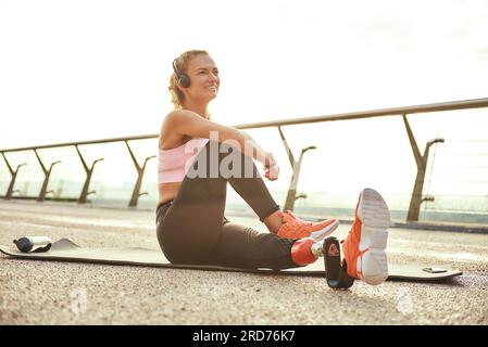 Buon giorno. Atleta portatrice di handicap positivo con protesi alle gambe in cuffie che fa esercizi di yoga e sorride mentre si siede sul ponte Foto Stock