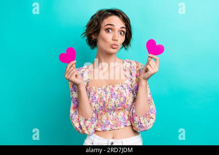 Ritratto di una camicetta con stampa floreale vestita da ragazza di buon umore con due cartoline a cuore rosa spedite a te un bacio isolato su sfondo color ottanio Foto Stock
