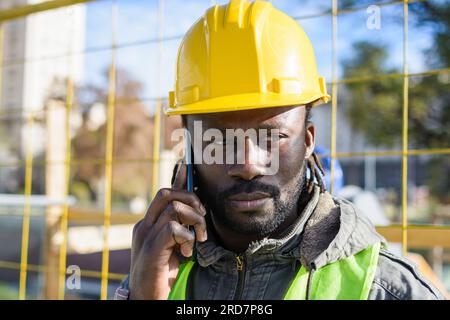 giovane dipendente nero del costruttore che indossa un elmetto di sicurezza giallo, in piedi all'aperto, ascoltando attentamente le istruzioni durante una telefonata. copia spazio. Foto Stock