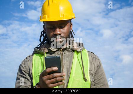giovane operaio edile di colore che indossa un elmetto di sicurezza giallo, una giacca e un giubbotto riflettente, mentre si trova all'aperto per leggere e inviare messaggi al telefono Foto Stock