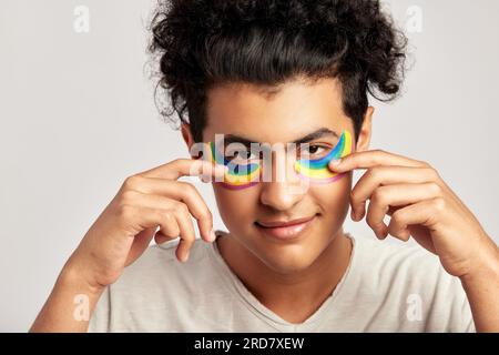 Ritratto di un giovane ragazzo bello con capelli ricci neri che applica macchie di idrogel di colore arcobaleno sotto gli occhi. Swarthy uomo sorridente che pratica la skincar Foto Stock