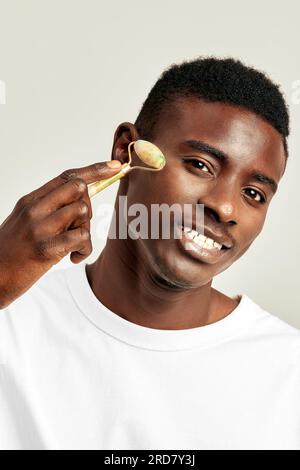 Un giovane ragazzo nero che usa il rullo facciale di degonfiore di giada per mantenere la sua pelle morbida e liscia. L'uomo millenario afroamericano mantiene la sua bea naturale Foto Stock