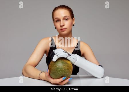 Giovane ragazza affascinante con disabilità che indossa protesi sensoriale bionica braccia sedute al tavolo. La bella donna tiene un melone nelle sue mani completamente sotto controllo Foto Stock