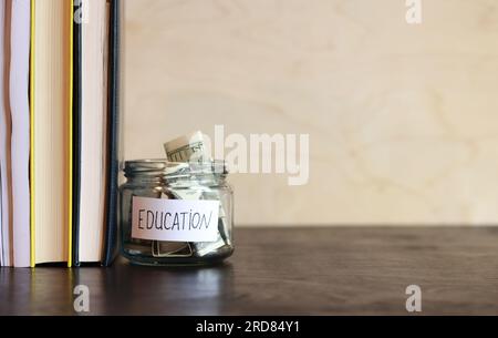 Vaso di vetro con denaro vicino a una pila di libri. Salvadanaio con dollari per l'istruzione su un tavolo di legno. Risparmia denaro per i futuri studi universitari. copia spac Foto Stock