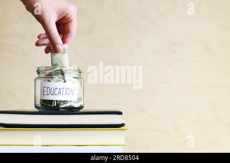 Salvadanaio con dollari per l'istruzione. Cumulo del concetto finanziario. Un vaso di vetro con soldi si trova su una pila di libri. La donna mette più soldi Foto Stock