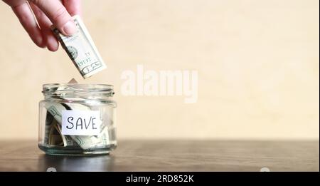 Risparmia denaro per il futuro. La mano di una donna mette una banconota da un dollaro in un barattolo di risparmi. Vaso di vetro con dollari su un tavolo di legno. Salvadanaio con banconote clo Foto Stock