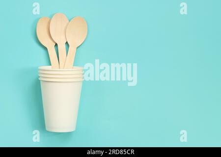 Cucchiai monouso in legno in una tazza di carta bianca, vista dall'alto. Utensili da cucina ecologici monouso su sfondo blu, spazio per la copia. Spianatura piatta. La conc Foto Stock