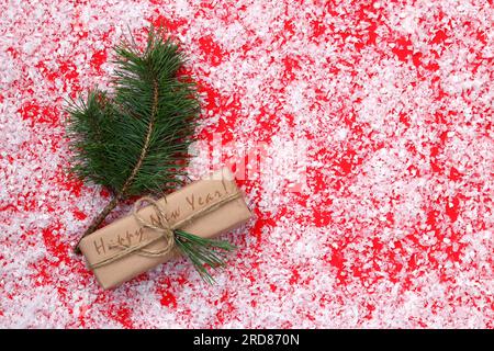 Un regalo per il nuovo anno vicino a un ramo di abete rosso, una composizione su sfondo rosso ricoperto di neve artificiale. Sfondo o copertina natalizia Foto Stock