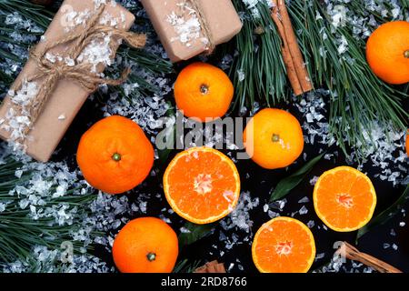 Composizione natalizia ricoperta di neve, sfondo con mandarini tagliati e interi, vista dall'alto. Agrumi, regali, rami di pino e cannella Foto Stock
