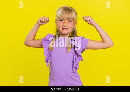 Sono forte e in salute. Giovane ragazza carina che mostra bicipiti e sembra fiduciosa sentendo la forza di lottare per i diritti, l'energia per ottenere il successo vince. Bambino biondo preteen su sfondo giallo Foto Stock