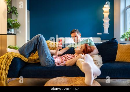 Coppia amorevole che riposa sul divano usando le app per smartphone. Moglie che mostra interessante sito web a marito Foto Stock