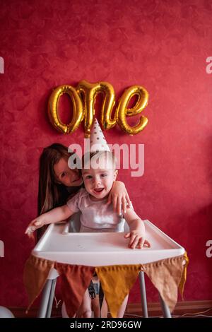 La sorella maggiore si congratula con il fratello minore per il suo primo compleanno. La ragazza bacia gli abbracci da bambino. I bambini si divertono su sfondo rosso con la lamina B. Foto Stock