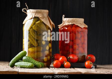 Cetrioli in scatola cetriolini e pomodori ciliegini in barattoli, verdure fresche per la conservazione su fondo di legno scuro. Foto Stock