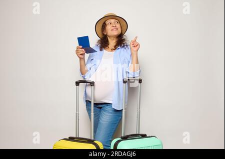Bella donna incinta con due valigie in policarbonato e carta d'imbarco, che va in vacanza all'estero, sorridendo a guardare da parte una copia dello spazio pubblicitario Foto Stock
