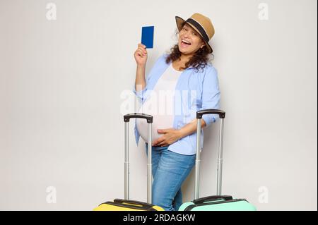 Felice femmina gravida multietnica che tocca la pancia incinta, sorride guardando la macchina fotografica, posa con carta d'imbarco, documenti di passaporto e due valigie Foto Stock