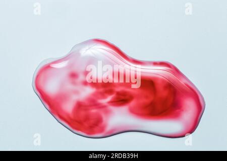 Goccia con acqua e sangue su sfondo blu. Goccia lucida colorata con riflesso sulla carta in primo piano. Sopra vernice cremisi traslucida spruzzata Foto Stock