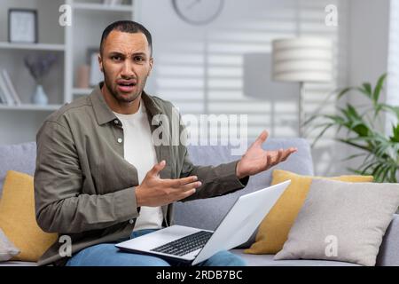 Giovane uomo d'affari afroamericano, libero professionista che lavora da casa con un portatile. Guarda con preoccupazione e rabbia la telecamera, gestendo inspiegabilmente con le mani. Foto Stock