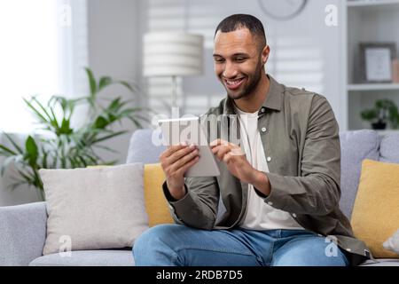 Giovane ispanico sorridente che usa un tablet a casa sul divano. Foto Stock