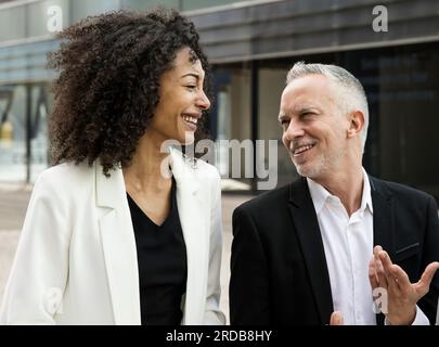 Donna d'affari e uomo d'affari che parlano e sorridono per strada. Incontro informale tra un giovane dirigente e un ceo senior che si diverte all'aperto. Foto Stock