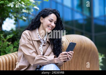 Bella donna allegra seduta sulla panchina fuori dall'edificio degli uffici, donna d'affari latinoamericana di successo che usa il telefono, legge notizie online e naviga nelle pagine online, tiene in mano lo smartphone. Foto Stock