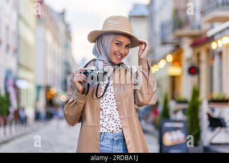 Ritratto della giovane bella donna musulmana in hijab e cappello, passeggiata turistica in città serale con macchina fotografica, donna che viaggia in diversi paesi. Foto Stock