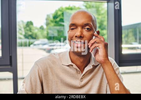 malattia di miastenia, uomo afro-americano felice al lavoro, allegro e scuro lavoratore di ufficio con la pelle di ptosi che parla su smartphone, inclusione, Foto Stock