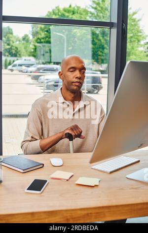 inclusione, audace uomo afro-americano con miastenia gravis, lavoratore d'ufficio dalla pelle scura seduto con bastone da passeggio e usando il computer, guardando la scimmia Foto Stock