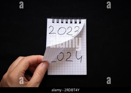 Blocco note con testo 2023 e 2024 e la mano gira la pagina del blocco note, Capodanno 2024 su sfondo nero, inizio di un nuovo anno Foto Stock