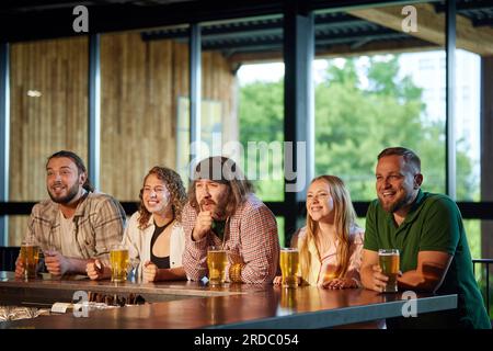 Gente allegra, amici seduti al bar, guardano con attenzione la traduzione delle partite di calcio, bevono, bevono birra, si divertono Foto Stock