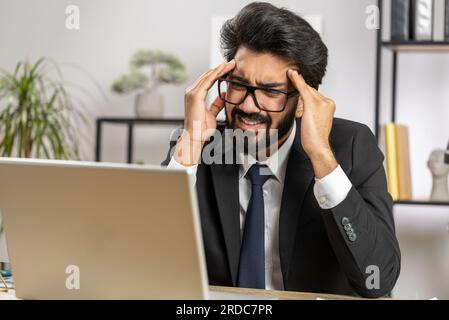 Uomo d'affari indiano malato stanco che soffre di mal di testa tensione problema ed emicrania, stress a casa scrivania ufficio posto di lavoro. L'uomo di lavoro di broker freelancer con bearded lavora su notebook. Lavoro eccessivo Foto Stock