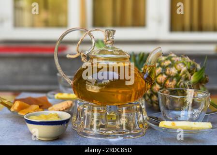 Teiera di vetro con tè all'ananas e marmellata di casa, tazze e frutta cristallizzata su sfondo scuro Foto Stock