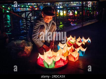 Un'anziana donna vietnamita siede su un ponte per vendere lanterne galleggianti con candele accese all'interno ai turisti di Hoi An, in Vietnam. Foto Stock