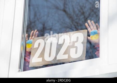 Una bambina tiene un cartone con il testo 2023 e la bandiera dell'Ucraina è dipinta sulle mani nella finestra della sua casa nel Dnieper in Ukrain Foto Stock