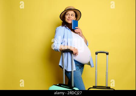 Una bellissima e deliziosa donna incinta multietnica tiene in mano il passaporto, sorride e penosamente guarda da parte uno spazio per le copie, potendo con le valigie sopra il giallo Foto Stock