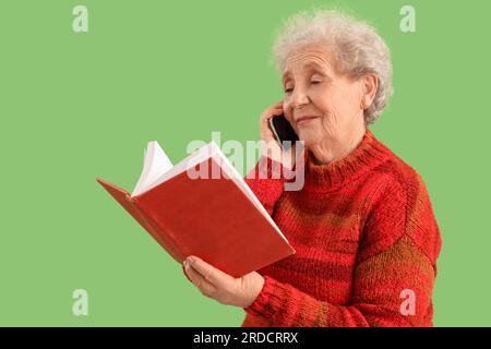 Donna anziana con un libro che parla da cellulare su sfondo verde Foto Stock