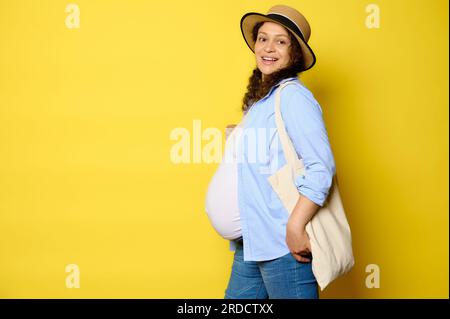 Ritratto di una giovane donna incinta multietnica che indossa un cappello di paglia e abiti casual in denim, che porta con sé una borsa per la spesa in lino ecologico, sorridente Looki Foto Stock