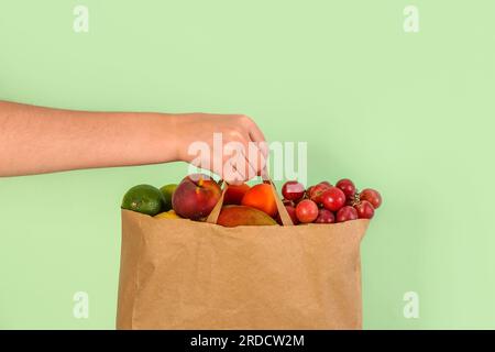 Mano femminile con sacchetto di carta pieno di frutta fresca su sfondo verde Foto Stock