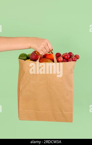 Mano femminile con sacchetto di carta pieno di frutta fresca su sfondo verde Foto Stock
