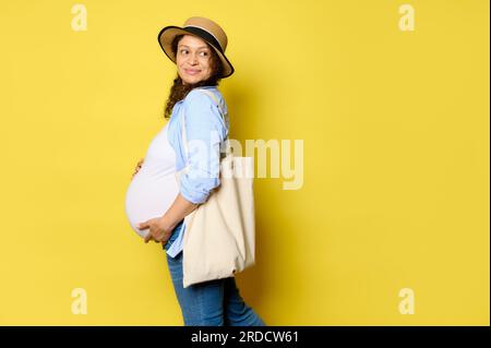 Autentica bella donna incinta riccia con cappello di paglia e abbigliamento casual, tiene la pancia, sorride graziosamente guardando da parte lo spazio per le copie, portando biancheria ecologica Foto Stock
