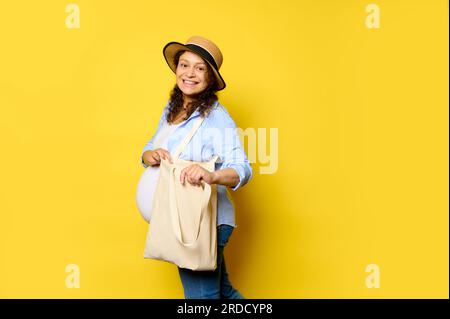 Una bella donna incinta multietnica sorride con un bel sorriso guardando la macchina fotografica, portando con sé una borsa per la spesa in lino ecologico su sfondo giallo isolato con annunci pubblicitari Foto Stock
